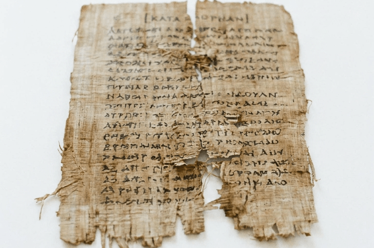Worn manuscript fragments laid out on a wooden table with ink pot, quill, and reading glasses